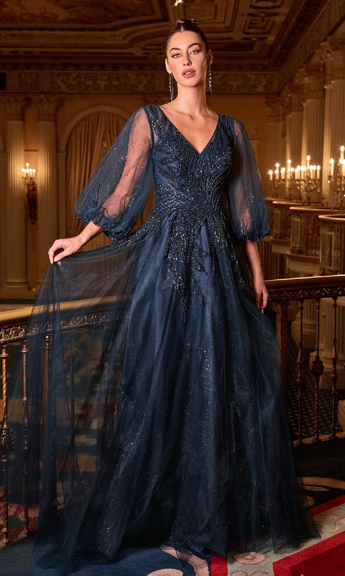 A woman stands indoors in the Ladivine CD0101 Balloon-Sleeve Blue Formal Gown, a navy evening dress with glitter details, voluminous sleeves, a V-neckline, and ornate decor. Chandeliers decorate the background.